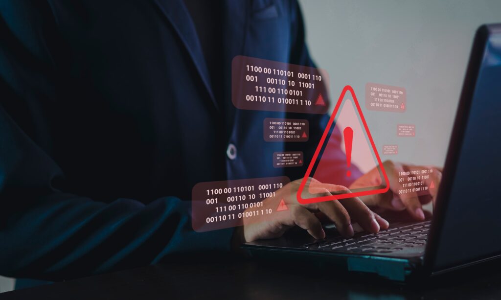 An individual working on their laptop with a futuristic hologram of a warning sign and IT coding in red